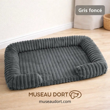 Lit pour chien orthopédique gris foncé avec rebords rassurants dans un salon, disponible chez Museau Dort